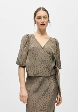 Kobieta ubrana w taupe'ową bluzkę z zakładką i dopasowaną spódnicę z subtelnym kwiatowym wzorem, stojąca na jasnym, jednolitym tle.