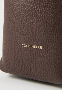 Brown leather bag with textured surface, featuring the embossed brand name "COCCINELLE" in gold on the side.