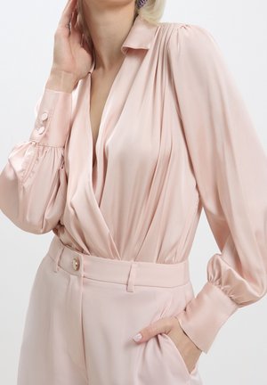 Femme portant un chemisier en satin rose pâle avec manches froncées et pantalon taille haute assorti, main dans la poche, sur un fond uni.