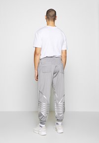 Muž stojící zády, oblečený v bílé tričko, šedých teplácích s reflexními pruhy na lýtkách, bílých teniskách a bílých ponožkách s černým pruhovaným vzorem.