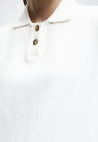 Λευκό πλεκτό πόλο με γιακά και δύο καφέ μαρμάρινα κουμπιά στο μπροστινό πέτο, που φοριέται από ένα άτομο.