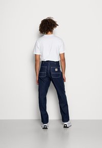 Pantalones de mezclilla azul oscuro con un corte relajado, costuras visibles y dos bolsillos traseros. Combinados con una camiseta blanca lisa y zapatillas.
