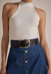 Witte geribbelde haltertop gecombineerd met een spijkerrok met gouden knoopaccenten en een brede zwarte leren riem met een gouden gesp.