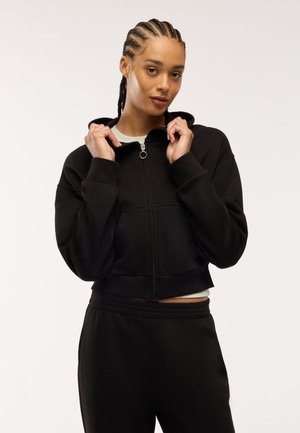 Donna con capelli intrecciati che indossa una felpa nera con zip e pantaloni neri, tenendo il colletto della felpa davanti a uno sfondo chiaro e uniforme.