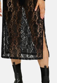 Fekete csipkeruha virágmintával, oldalán nyílásokkal. Testhez simuló fazon, texturált felülettel és fodros szegéllyel. Látható fekete csizma.