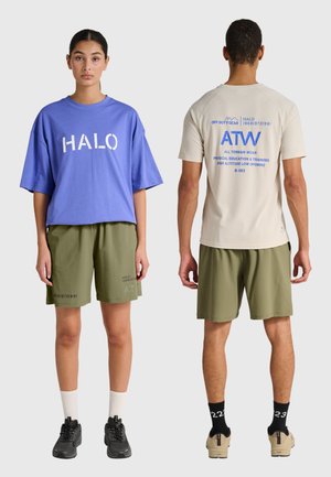 Two models wearing casual athletic outfits: one facing forward in a blue oversized shirt and olive shorts, the other facing back in a beige shirt with blue text.