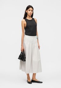 Γυναίκα με μαύρο αμάνικο τοπ και λευκή πλισέ μίντι φούστα, κρατώντας μια μικρή μαύρη τσάντα, φορώντας μαύρες μπαλαρίνες, στέκεται πάνω σε λευκό φόντο.