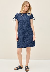 Tummansininen mekko, jossa on lehtikuosi, lyhyet hiha- ja nauhakiinnitykset sekä rento malli, yhdistettynä tummaan sandaaliin vaalealla taustalla.
