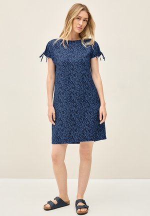 Dunkelblaues Kleid mit einem Blattmuster, kurzen Ärmeln mit Bindebändern und relaxter Passform, kombiniert mit dunklen Sandalen auf hellem Hintergrund.