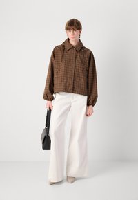 Braun karierte kurze Jacke mit Kordelzugkapuze, kombiniert mit weißen weit geschnittenen Hosen. Das Modell hält eine schwarze Handtasche. Glatte Textur.