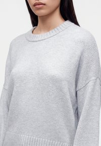 Jeune personne aux longs cheveux foncés portant un pull en tricot gris clair avec un col et des poignets côtelés.
