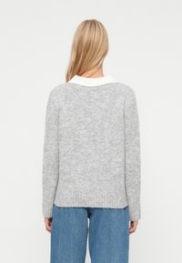 Blonda sieviete zilos džinsos, uzvilkusi gaiši pelēku adītu džemperi pāri baltai krekla apkaklei, rādīta no aizmugures uz viendabīga fona.