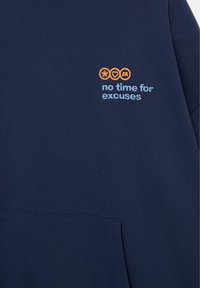 Material bleu închis cu text brodat în albastru deschis „fără timp pentru scuze” sub trei cercuri portocalii conținând o floare, o inimă și o literă R stilizată.