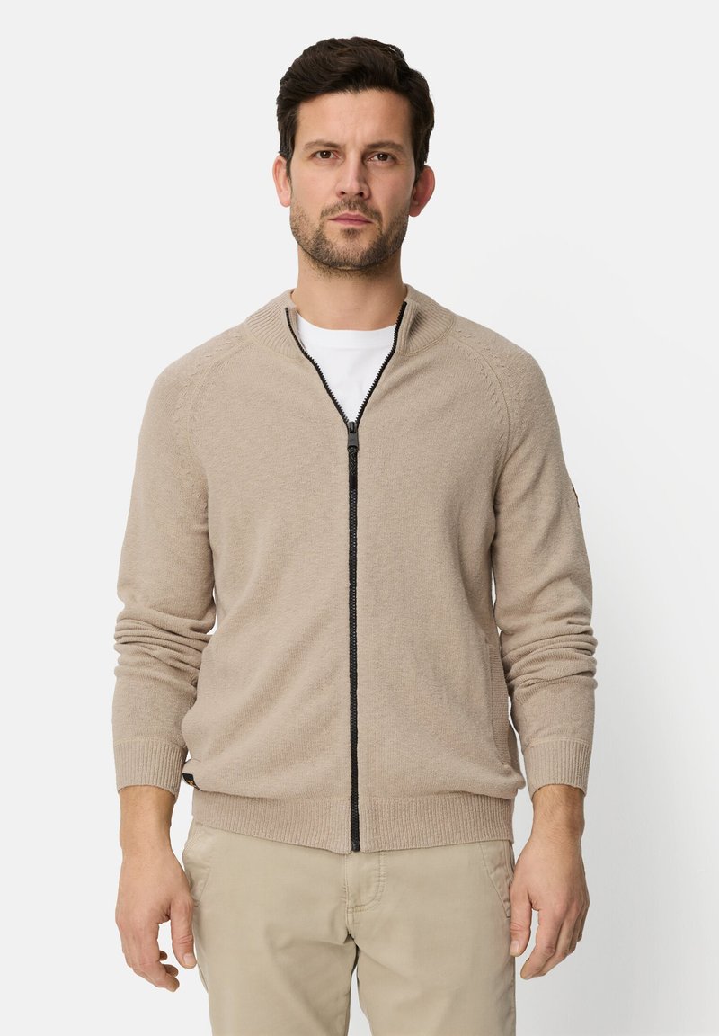 Beige zip-up cardigan made of soft knit material, featuring a ribbed collar and cuffs, with a black zipper, worn over a white T-shirt.