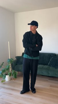 Fekete, túlméretezett dzseki türkizkék inggel, széles fekete nadrág és fekete cipő. Az összeállítást egy tengerészkék sapka teszi teljessé. A háttérben egy zöld kanapé látható.