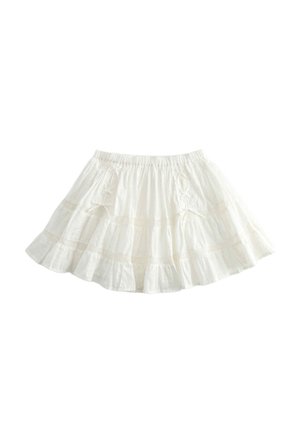 Giovane donna con capelli lunghi e ricci che indossa una giacca bianca con cerniera, una gonna bianca a balze, calzini beige e scarpe marroni con lacci rossi.