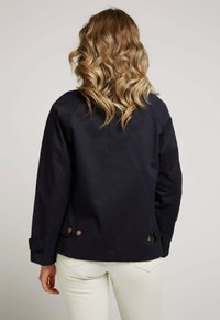 Veste bleu marine au design décontracté, dotée de deux boutons marron à l'arrière et d'une coupe décontractée. Le tissu semble lisse avec une finition mate.