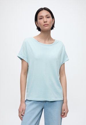 Νεαρή γυναίκα με κοντά μαύρα μαλλιά που φοράει ανοιχτό μπλε κοντομάνικο πουκάμισο και ανοιχτό μπλε τζιν, σε απλό λευκό φόντο.