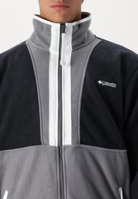 Grå och svart fleecejacka med vit dragkedja och randdetalj. Har en hög krage och Columbia-logotypen på bröstet.