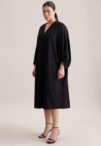 Robe noire ample avec un col en V et des manches ballon. Tissu lisse, longueur au genou, portée avec des sandales noires à lanières.