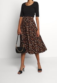 En kvinna poserar i en svart topp och en leopardmönstrad midikjol, håller en svart väska med en guldkedja. Hon har på sig svarta  öppetåade klackskor.