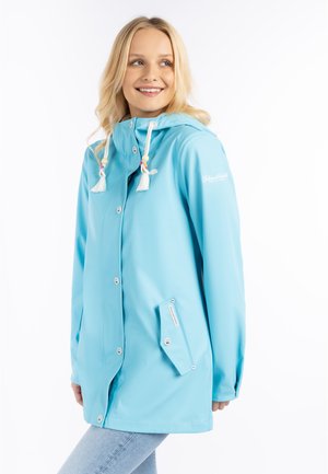 Glückliche Frau mit hellblauer Regenjacke mit Kapuze und weißen Kordeln, die seitlich vor einem weißen Hintergrund schaut.