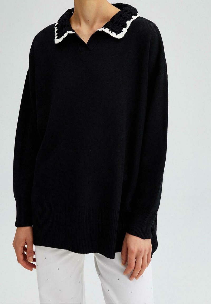 Femme portant un pull en maille noire ample avec un col festonné noir et blanc, associé à un pantalon blanc orné de petites décorations.