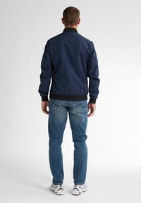Marineblauer Bomberjacke mit schwarzem Kragen, gerippten Bündchen und Saum. Kombiniert mit hellblauen Jeans und weißen Turnschuhen, von hinten betrachtet.