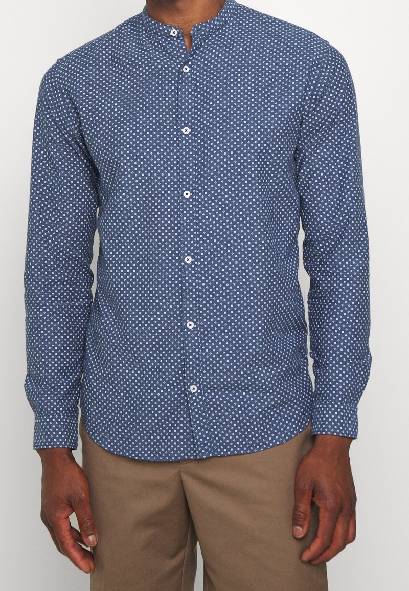 Mies, joka on pukeutunut siniseen istuvaan nappipaitaan, jossa on valkoisia geometrisia kuvioita, ja beigen värisiin housuihin, seisoo yksinkertaista taustaa vasten.