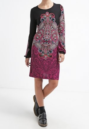 Kobieta w długim rękawie sukience w czarno-różowy wzór paisley, w połączeniu z czarnymi skarpetkami i czarnymi butami na sprzączkę, stojąca ze skrzyżowanymi nogami.