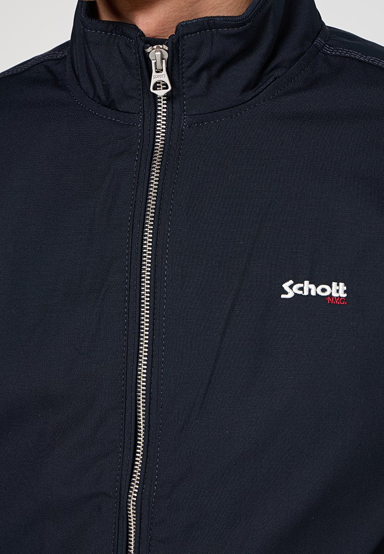 Veste zippée bleu marine en tissu lisse, dotée d'un col montant, d'une fermeture éclair argentée et d'un petit logo brodé en blanc et rouge sur la poitrine.