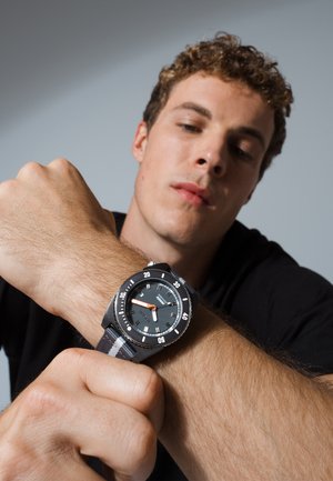 Man adjusts black wristwatch with gray-striped fabric band, focusing on the watch face showing time and minute markers against a plain background.