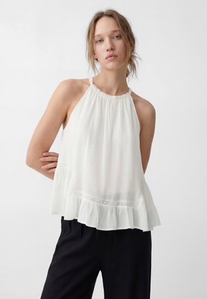 Junge Frau mit hellem Haar, die ein ärmelloses weißes Neckholder-Top mit Rüschensaum und schwarze Hose vor schlichtem Hintergrund trägt.