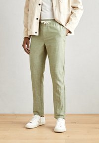 Light green, linen blend pants with elastic waistband, drawstring, and side seams. Paired with white sneakers on a wooden floor.