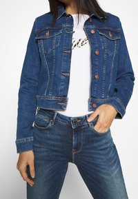 Džínová bunda v tmavě modré barvě s měděnými knoflíky, zkráceným střihem a dvěma náprsními kapsami. Nasažena přes bílé tričko se zlatým potiskem.