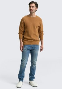 Suéter de cuello redondo en marrón claro, con textura acanalada, usado con jeans azul claro y zapatillas blancas. Hombre de pie con cabello corto oscuro, fondo neutral.