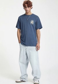 Blå bomullst-shirt med korta ärmar och en grafisk design på bröstet, kombinerad med ljusblå baggyjeans och sneakers.