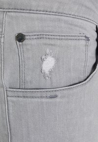 Ljust grå denimtyg har en bakficka med ett slitet hål och en metallknappdetalj som framhäver textur och slitage.