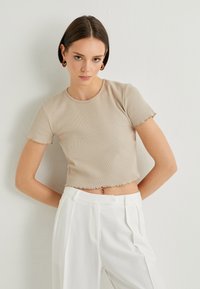 Top corto de canalé beige con mangas cortas y detalles de dobladillo sin terminar, combinado con pantalones blancos de talle alto. Textura suave, diseño ajustado.