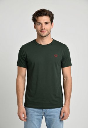 Giovane uomo che indossa una t-shirt verde scuro a maniche corte con un logo rosso sul petto e jeans azzurri, in piedi davanti a uno sfondo grigio semplice.
