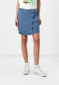 Denim obleka z zavihanimi robovi, modre barve, z gumbom za zapiranje na strani, v kombinaciji z belimi športnimi copati. Teksturna bombažna tkanina.