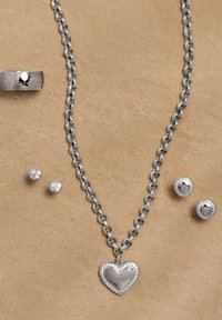 Conjunto de joyas de plata que incluye un collar con cadena y colgante en forma de corazón, pendientes de botón a juego y anillo grabado, todos con texturas brillantes.