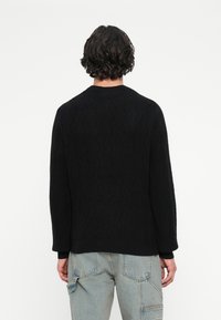 Muž s kučeravými tmavými vlasmi, oblečený v čiernom textúrovanom svetri a svetlomodrých džínsoch s cargo strihom, zobrazený zozadu na bielom pozadí.