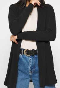 Schwarze Strickjacke mit offenem Vorderteil, gerippter Textur, langen Ärmeln, kombiniert mit einem beigen Oberteil und hochtaillierten blauen Jeans.