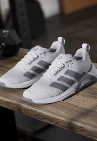 Paire de baskets blanches et grises avec lacets posées sur une table en bois, équipement de gym flou en arrière-plan.