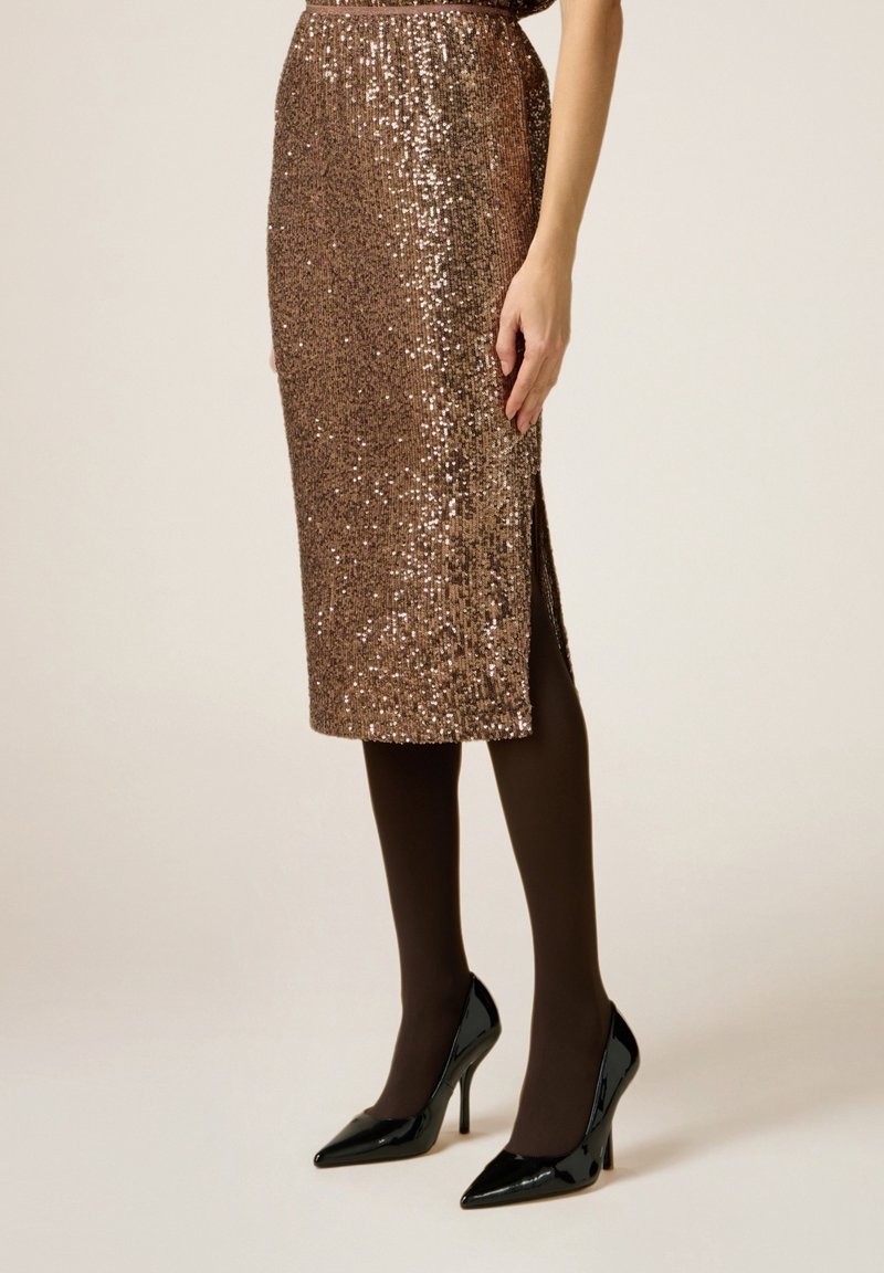 Femme portant une jupe marron à sequins longueur genou avec une fente latérale, des collants noirs et des chaussures noires à talons aiguilles pointues, debout sur un fond uni.