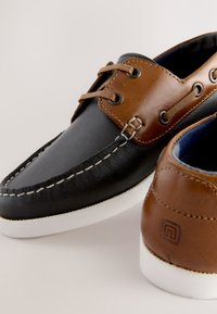 Chaussures bateau en cuir avec embout noir, détails en cuir marron, surpiqûres blanches et semelle blanche. Comprend un laçage à l'avant et un détail de logo.