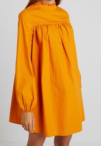 Oranssi, pitkähihaiset mekko kevyestä materiaalista, jossa on korkea kaulus, rypytetty rintamusta ja väljä istuvuus. Varustettu joustavilla hihansuilla ja minimaalisella muotoilulla.