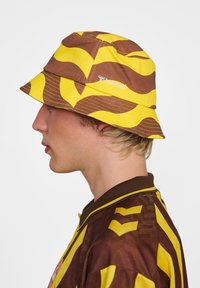Gul och brun mönstrad bucket hat med bred brätte, med en logotyp på sidan. Tillverkad av bomull med en mjuk textur.
