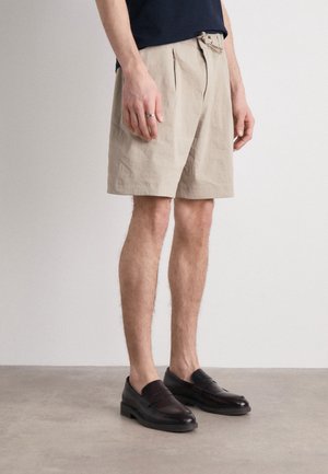 Pantaloni corti in cotone beige con pieghe, vita con cintura e tasche laterali, abbinati a mocassini in pelle marrone scuro su una superficie grigia chiara.